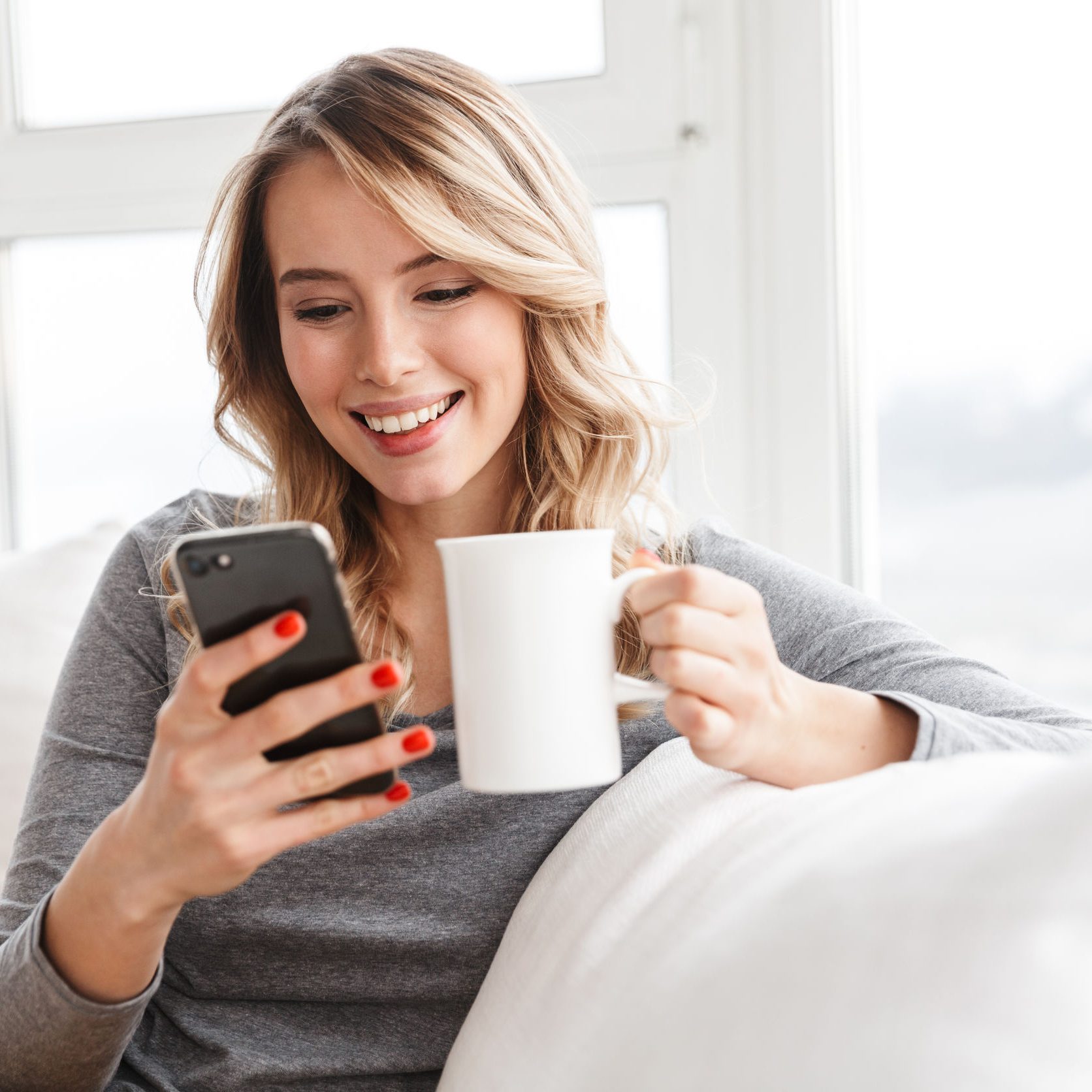 Image of cute pretty woman sitting indoors at home holding cup drinking tea using mobile phone.